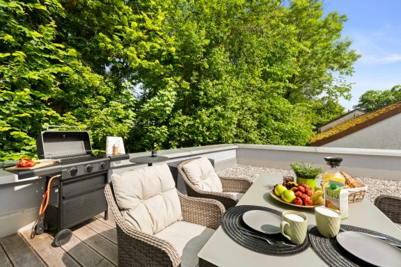 Ferienwohnung Miesbach – Flussperle: Terrasse mit Grill und einladendem Essbereich Terrasse mit Grill, gemütlichen Stühlen und gedecktem Tisch mit Obst und Getränken.