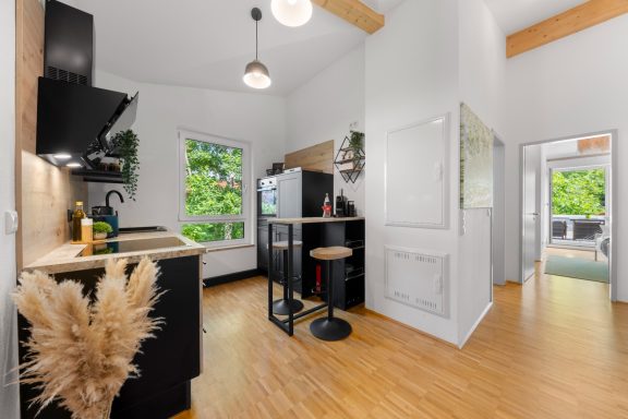 Ferienwohnung Miesbach – Flussperle: Moderne Küche mit Flussblick und stilvollen Holzdetails Moderne Küche mit Holzdetails, Barstuhl und großen Fenstern zum Außenbereich.