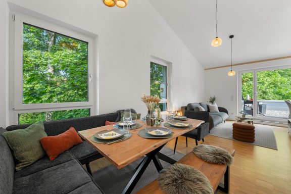 Ferienwohnung Miesbach – Flussperle: Helles Wohnzimmer mit Naturblick und Esstisch Helles Wohnzimmer mit einem Esstisch, Couch und großen Fenstern, umgeben von Bäumen.