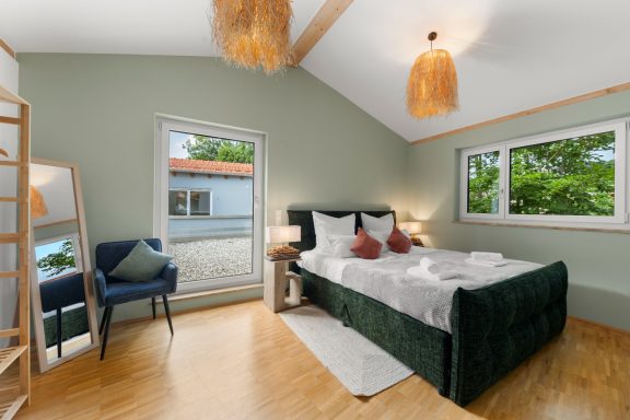 Ferienwohnung Miesbach – Liebelei: Helles Schlafzimmer mit grünem Bett und Holzdetails Helles Schlafzimmer mit einem grünen Bett, Fenster und Holzdetails.
